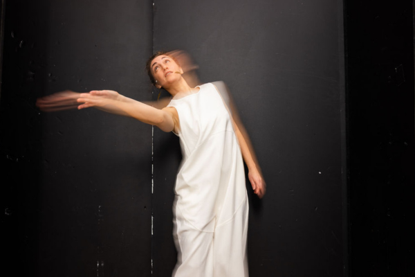 Woman in white gown moves in front of black background, blurred by camera exposure